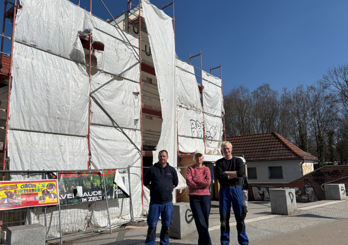 Künstlerin Barbara Trautmann und Mitarbeiter der Werkstatt für Metallgestaltung "Fittkau" stehen vor dem Bahnhof Hohen Neuendorf.